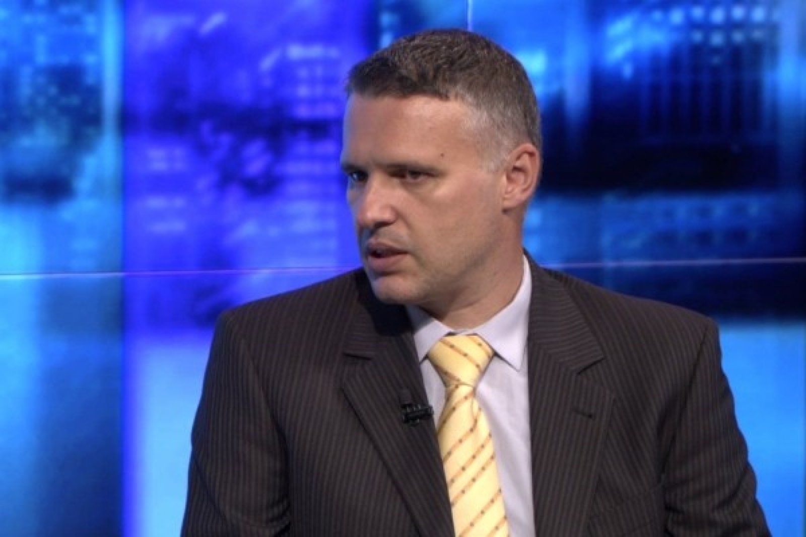 Man in striped suit and yellow tie speaking during a TV interview, with a blue blurred background.