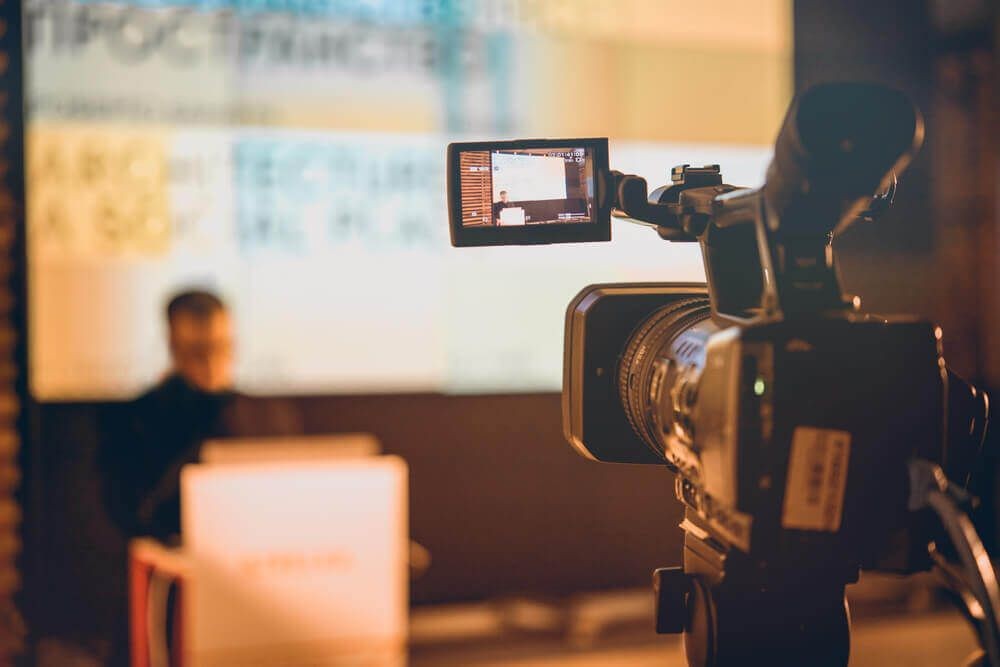 Close-up of a video camera recording a presentation with blurred speaker and screen in the background.